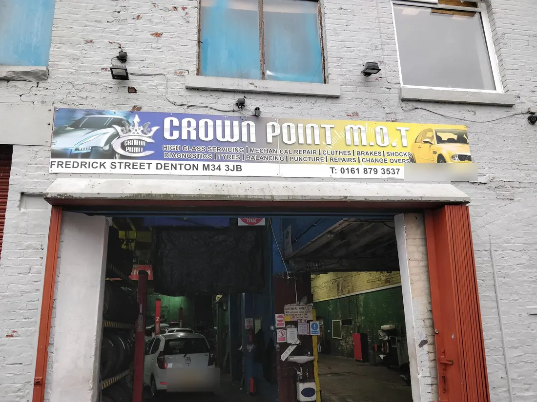 The front of Crown Point MOT Centre: a white-painted brick wall, the blue and yellow CROWN POINT M.O.T sign with its crown, and the open doorway into the workshop with a white car inside