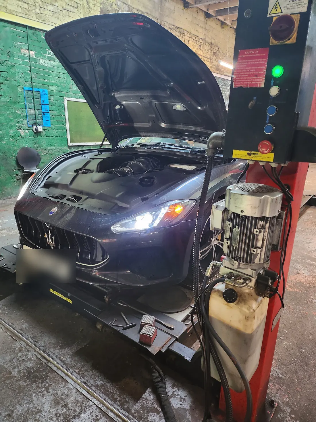 A dark Maserati with its bonnet up and headlights on, beside a red lift post