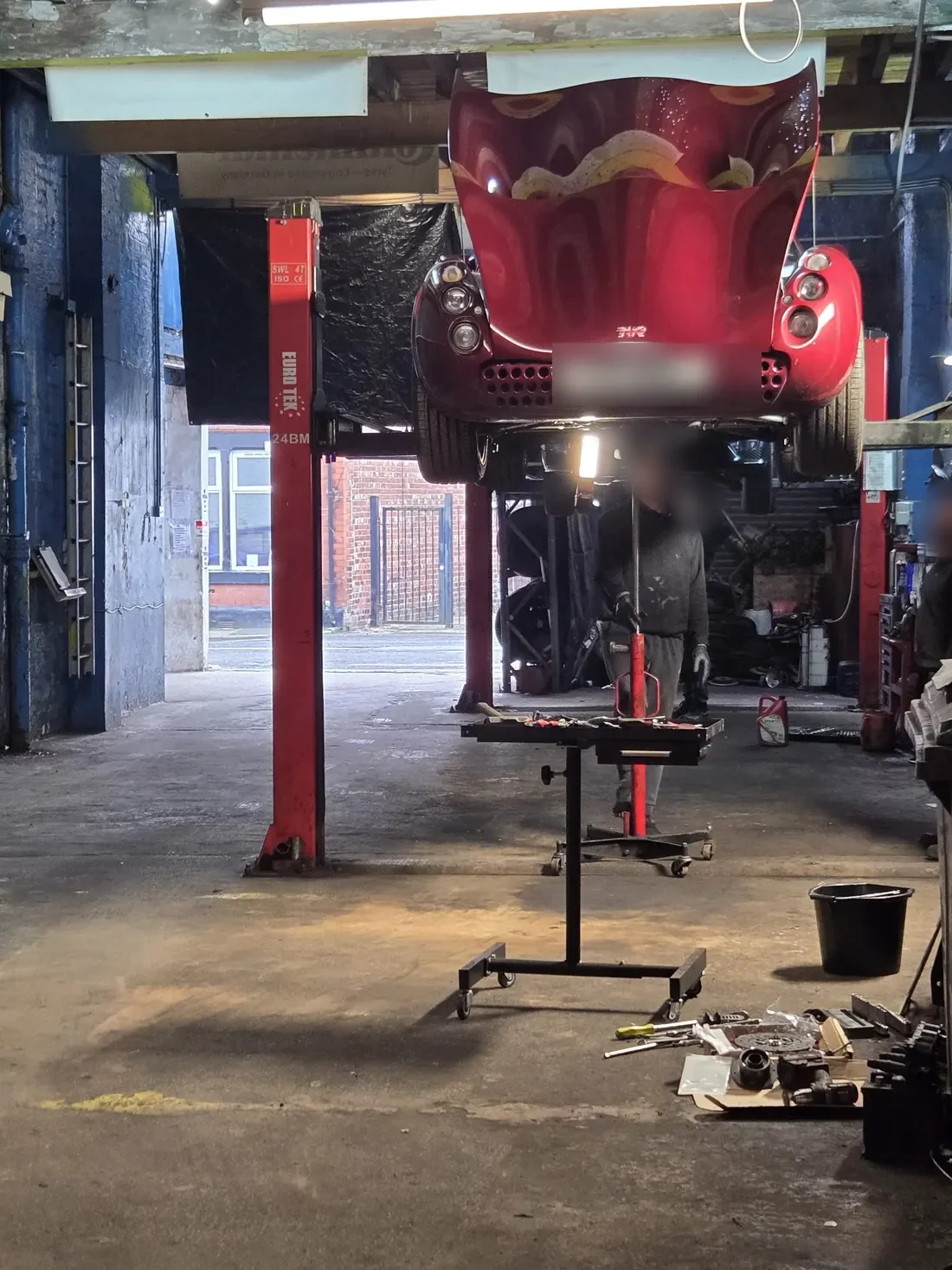 A red car high on the lift, a mechanic working underneath and a transmission jack below